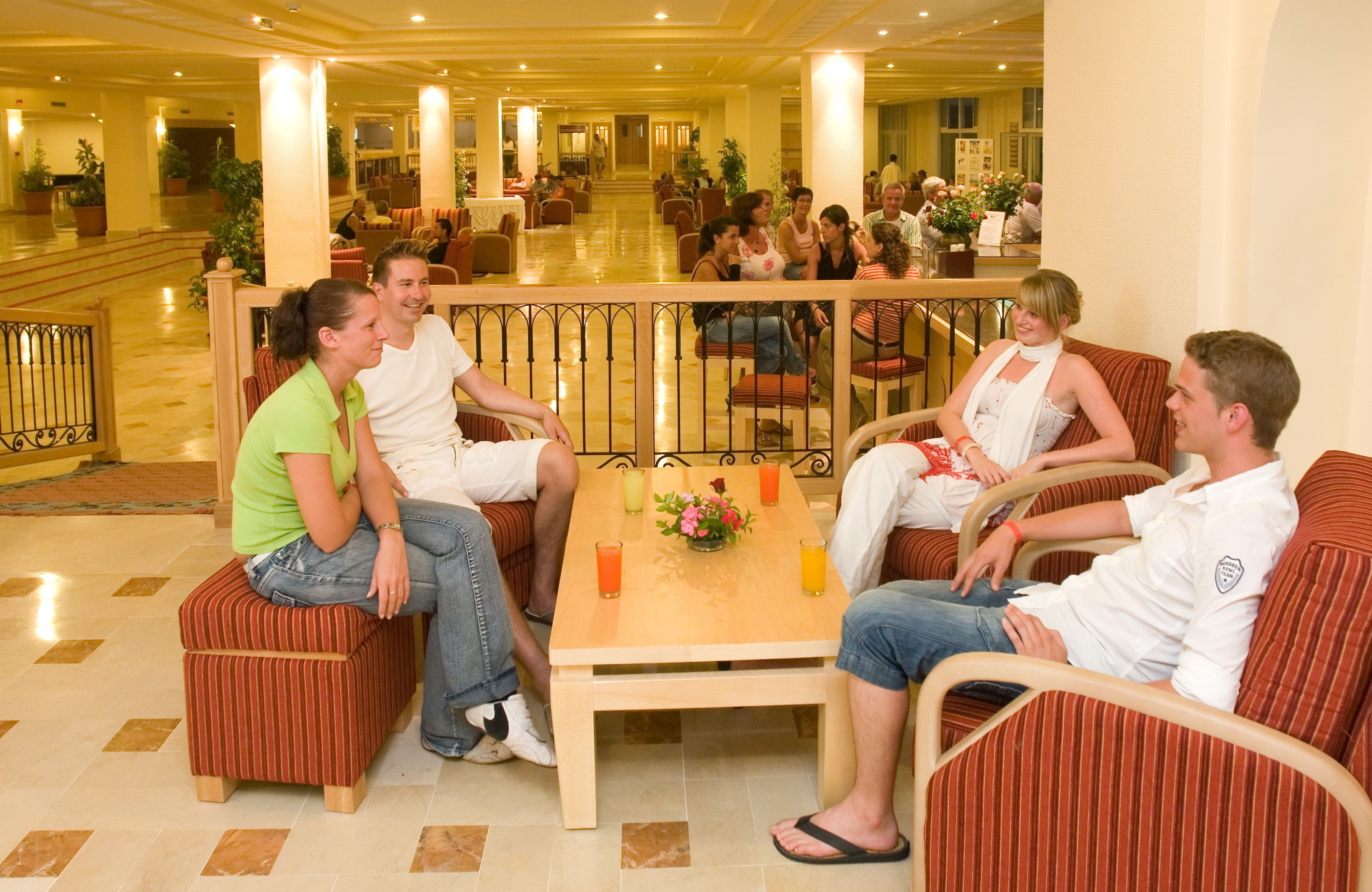 A group of people seated on cushioned furniture in a spacious, well-lit lobby with a tiled floor and a central table with drinks and a flower arrangement.