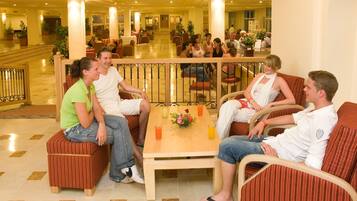 A group of people seated on cushioned furniture in a spacious, well-lit lobby with a tiled floor and a central table with drinks and a flower arrangement.