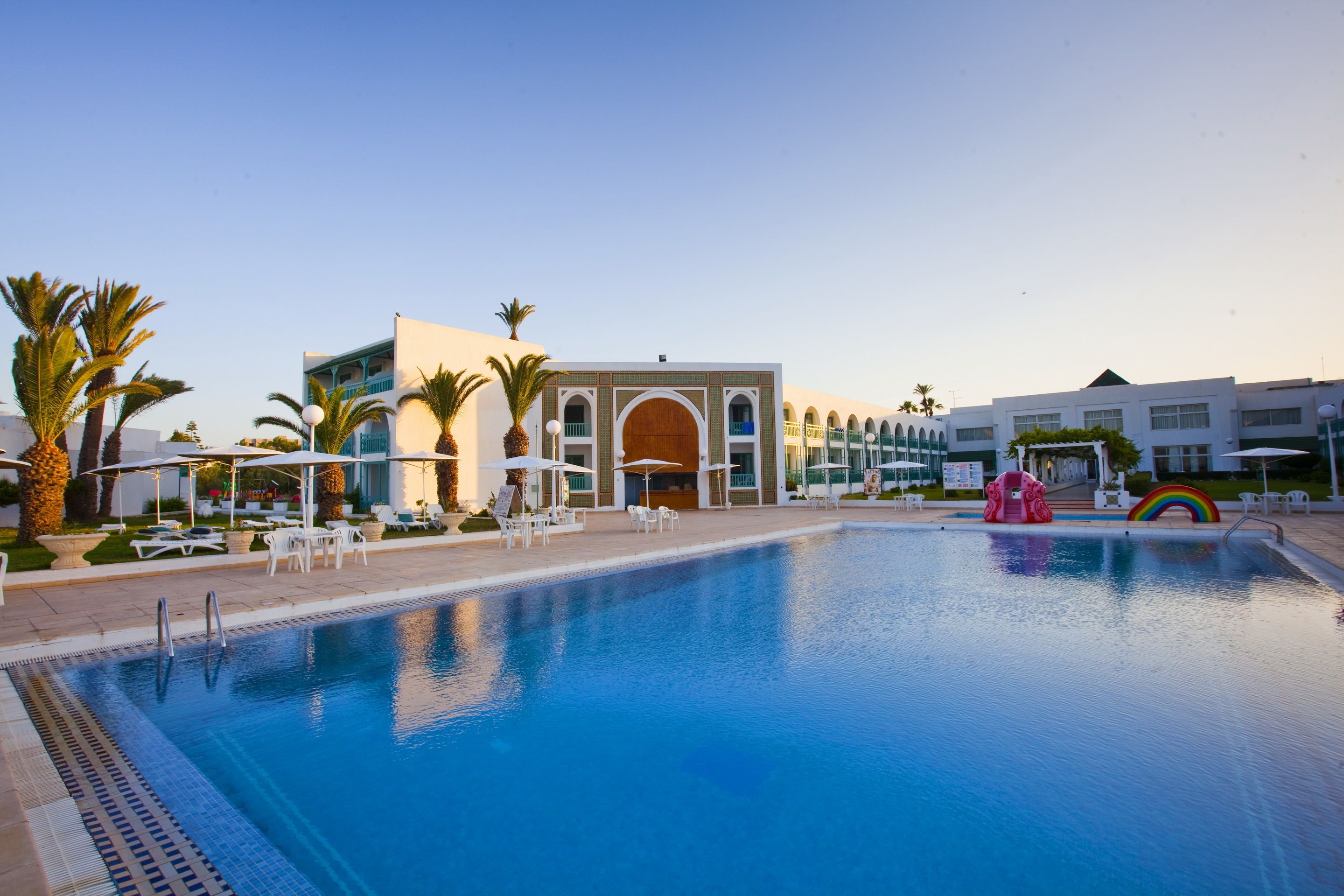 A swimming pool with lounge chairs and umbrellas, surrounded by palm trees and a building with arched windows.