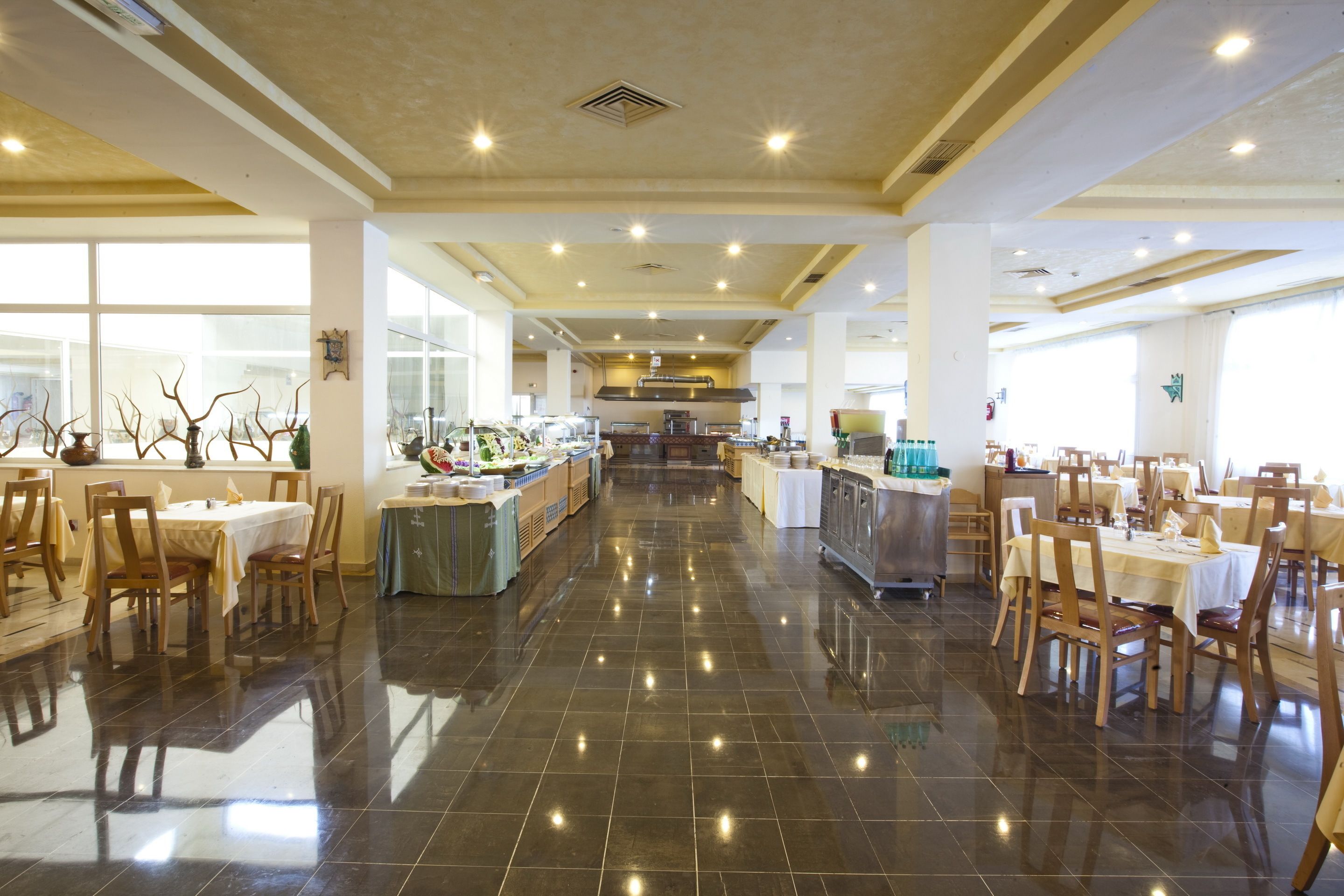 Amplio comedor de hotel con múltiples mesas y sillas, una cocina central y grandes ventanales que proporcionan luz natural.