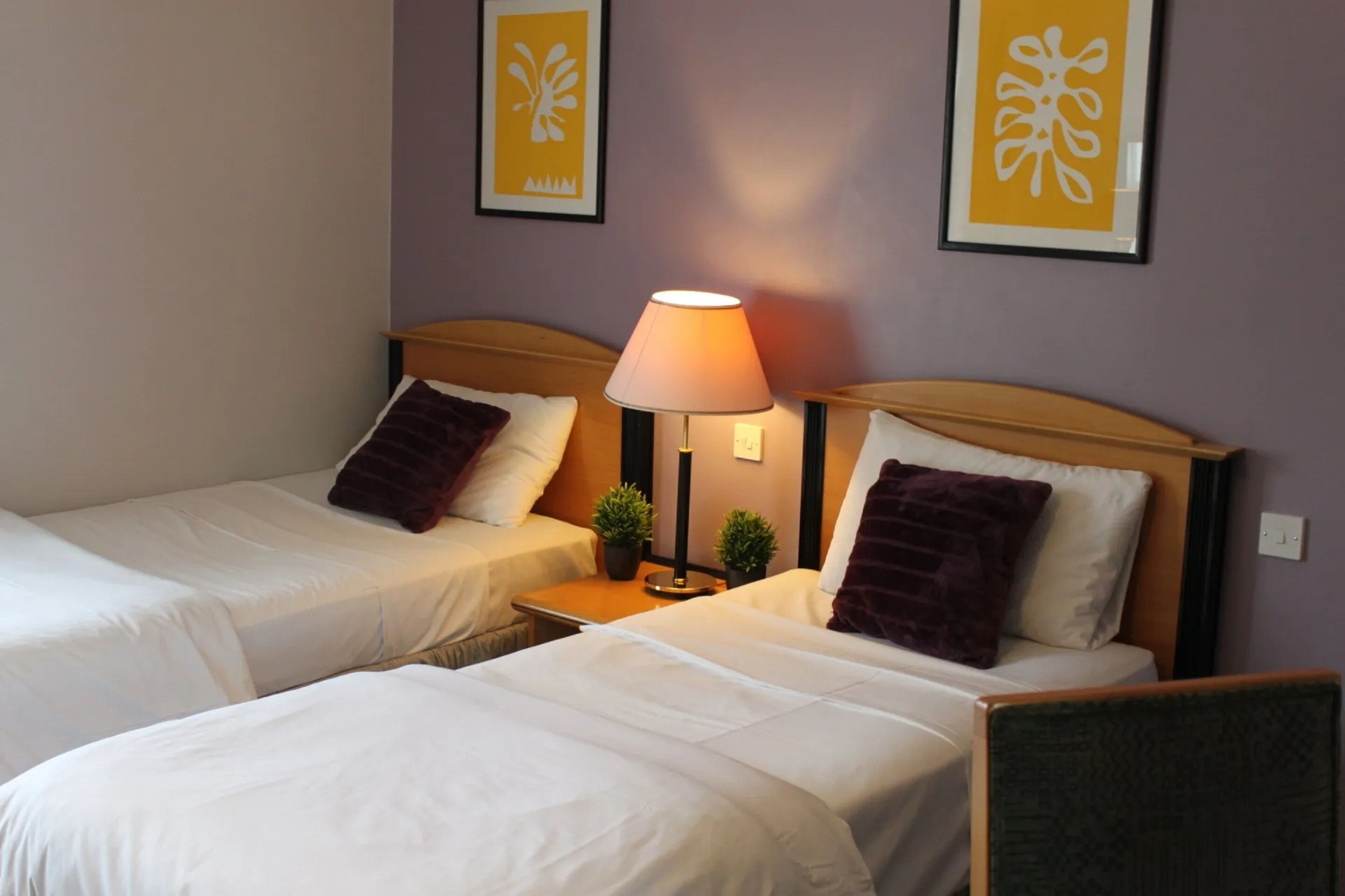 A hotel room with two single beds, a nightstand with a lamp, and two framed wall art pieces.