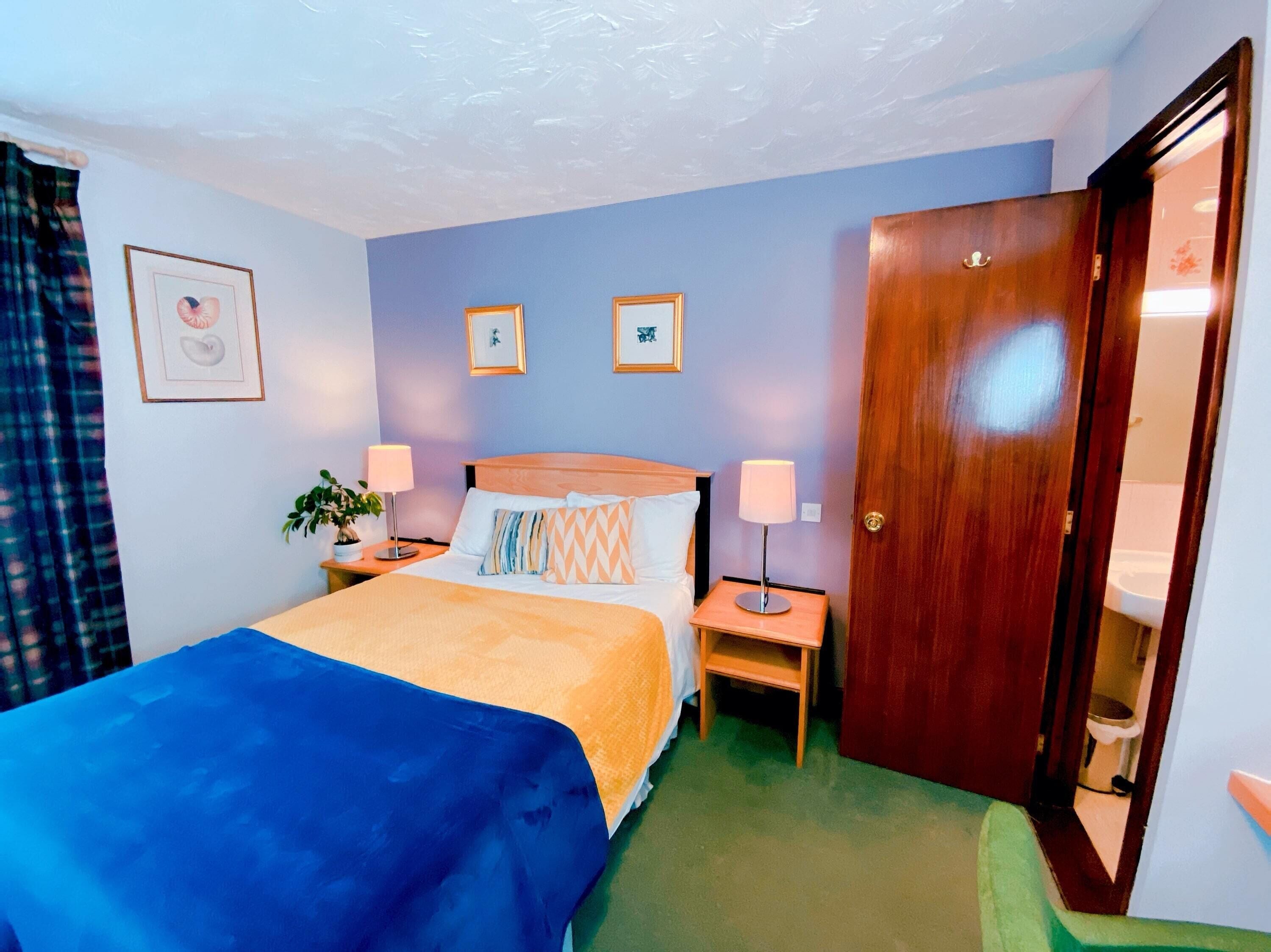 A hotel room with a bed, two bedside tables, a nightstand, a door, and a wall with two framed pictures.