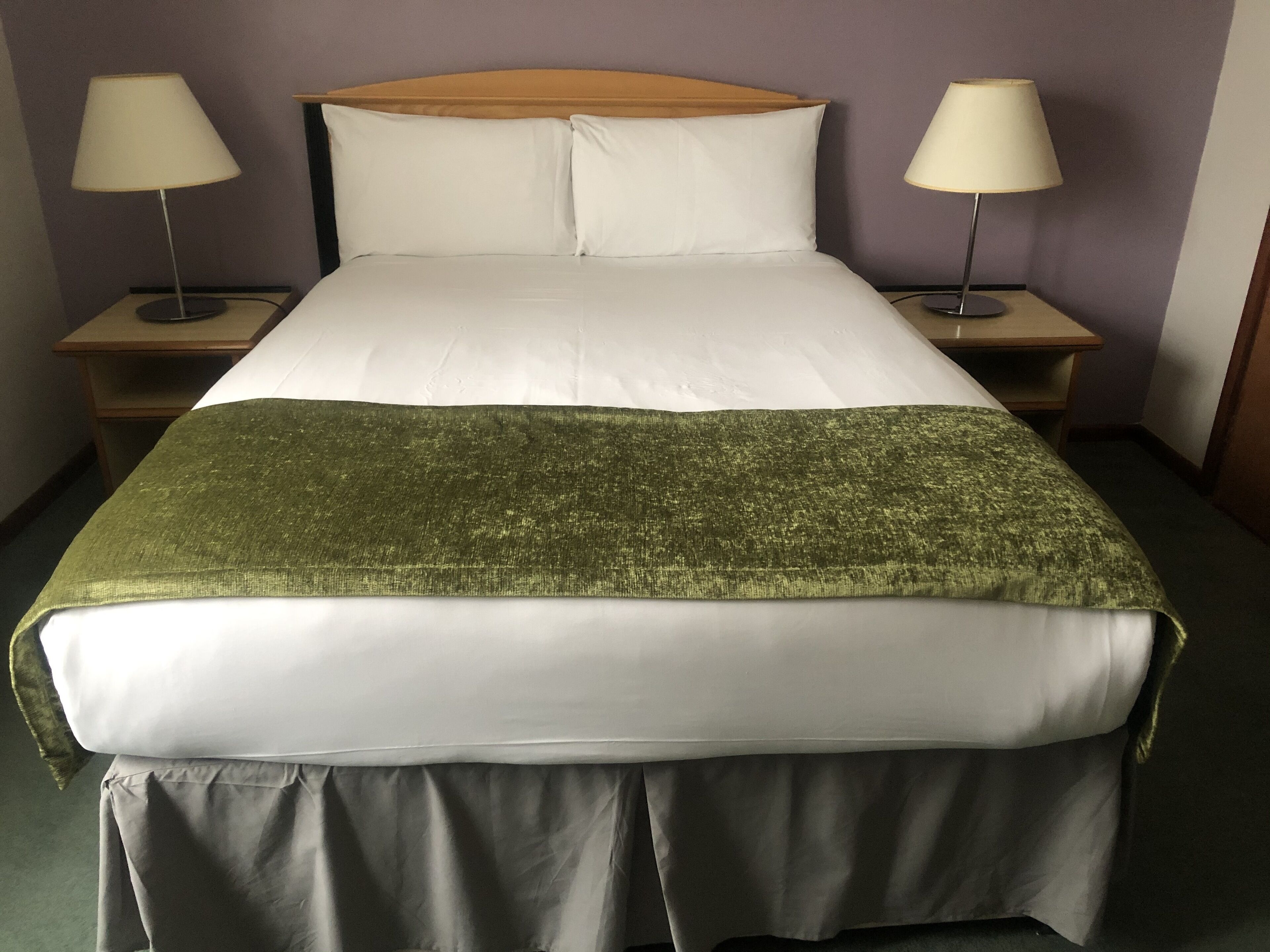 Una cama bien tendida con sábanas blancas, un plaid verde y un cabecero de madera. Hay dos lámparas de noche sobre mesitas de madera.