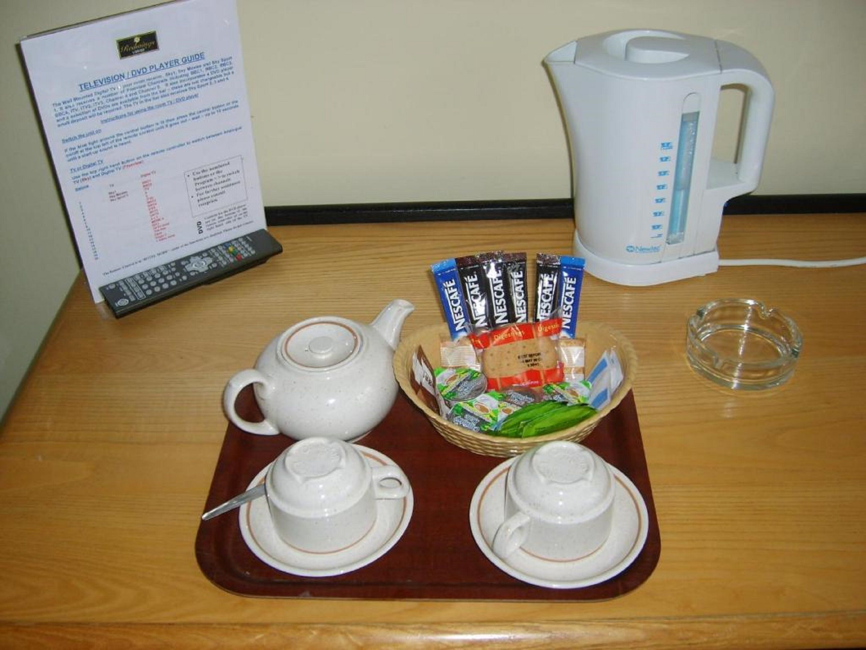 Una mesa con tetera, tazas, cesta de comida, hervidor y control remoto.