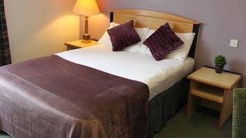 A neatly made bed with a brown bedspread, two purple pillows, and a wooden headboard. There is a nightstand with a lamp and a small potted plant beside the bed.