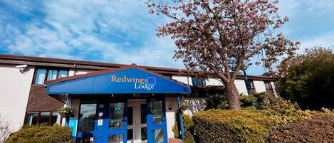 Exterior del Redwings Lodge con cielo azul despejado, una entrada acogedora y setos bien cuidados.