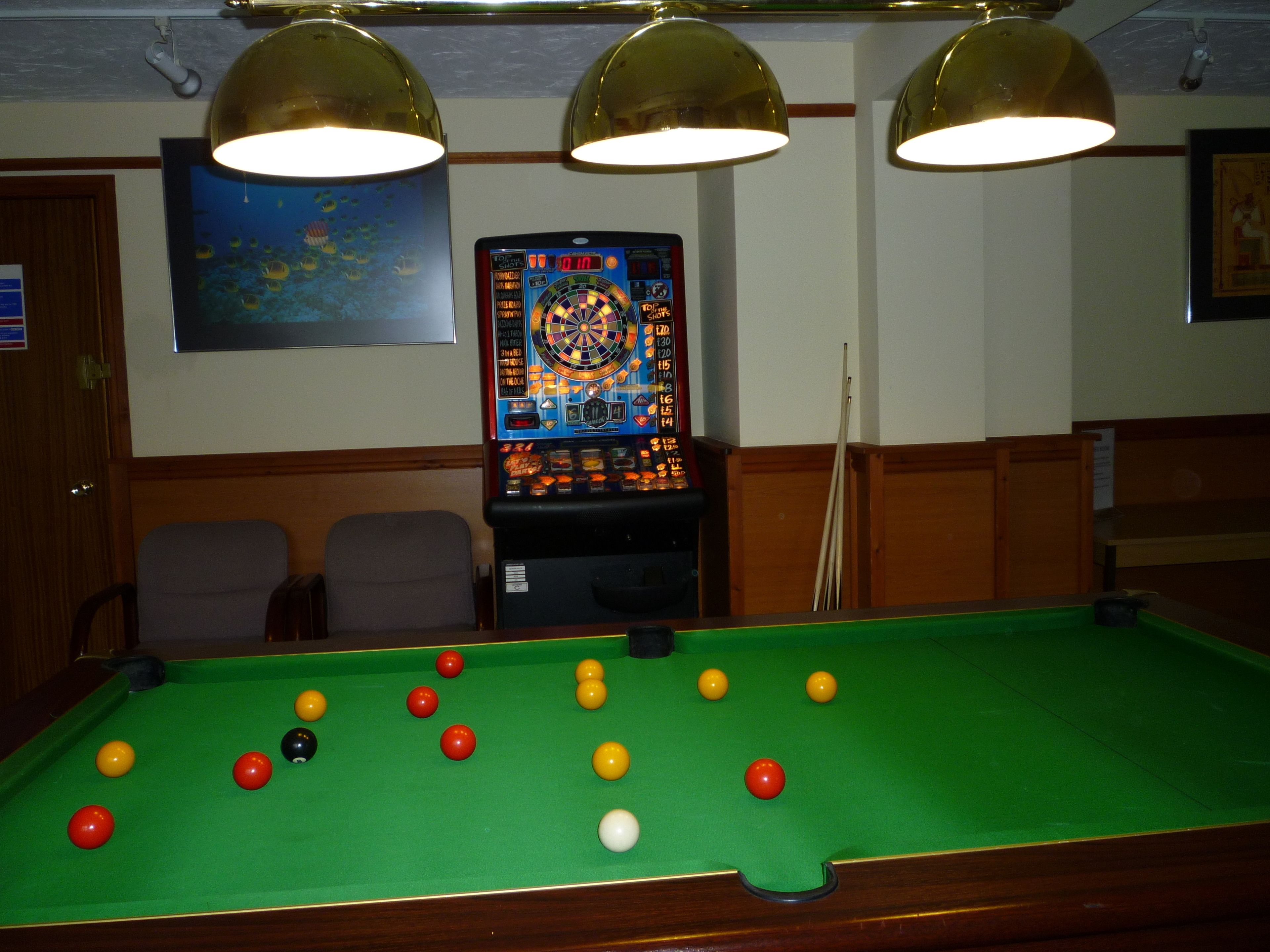 Ett biljardbord med bollar, en darttavla och taklampor i ett rum med träpanel och en inramad tavla på väggen.