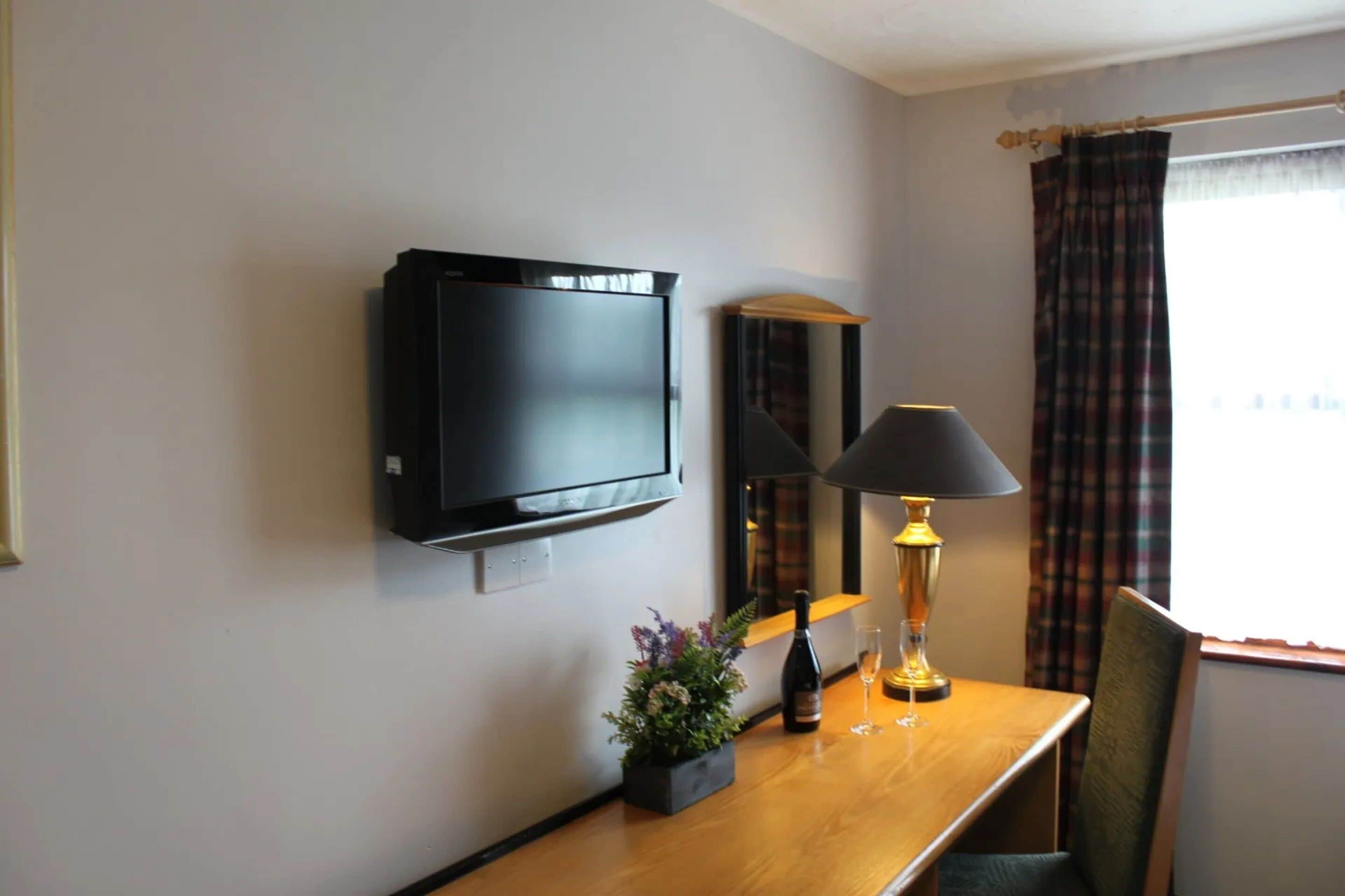 Et hotelværelse med et vægmonteret fladskærms-tv, et træskrivebord med lampe, en lille plantet potte og en flaske vin.