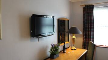 Une chambre d’hôtel équipée d’un téléviseur à écran plat fixé au mur, d’un bureau en bois avec une lampe, d’une petite plante en pot et d’une bouteille de vin.