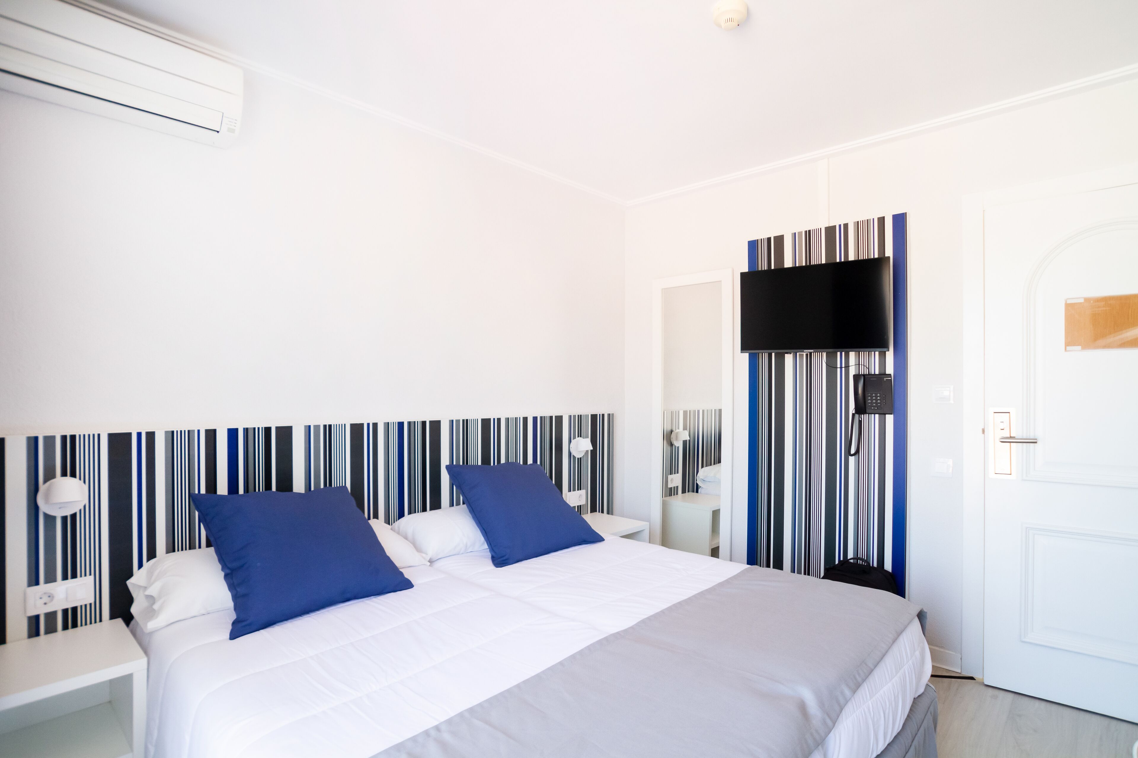 Een hotelkamer met een bed, witte muren, een gestreept hoofdeinde, een flatscreen televisie en een deur.