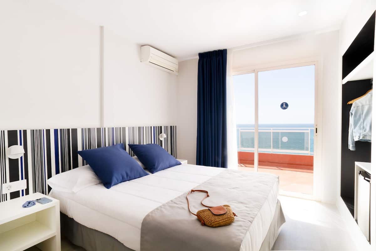 A hotel room with a bed, a small bedside table, a window with a view of the sea, and a balcony with a railing.