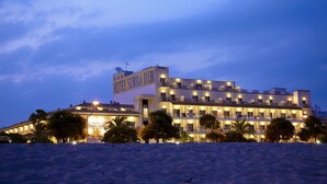 Un hotel de varios pisos con un cartel llamativo que dice "HOTEL SORRA D'OR" al anochecer. El edificio tiene múltiples balcones y está rodeado de palmeras.