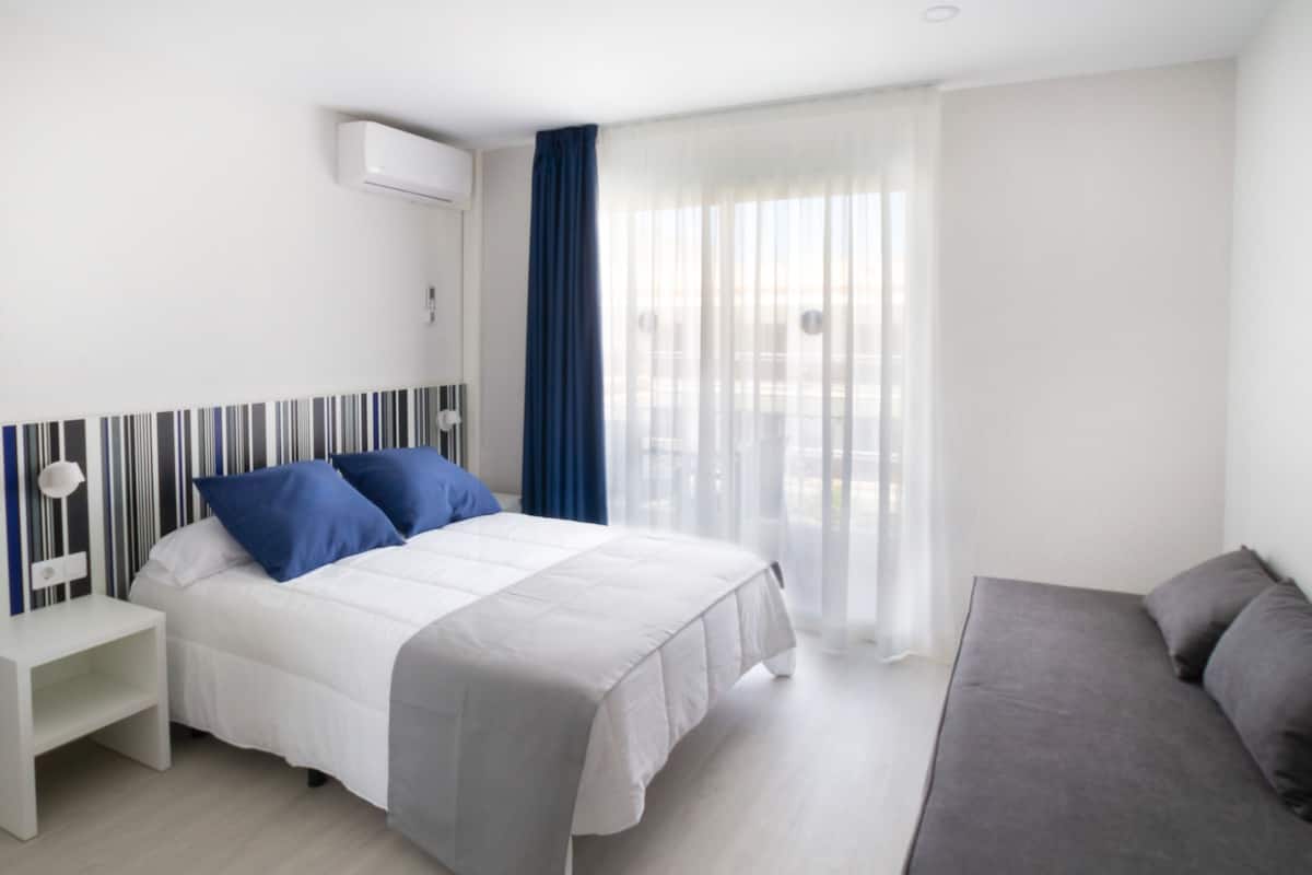 A modern hotel room with a bed, a sofa, a nightstand, and a window with sheer curtains.