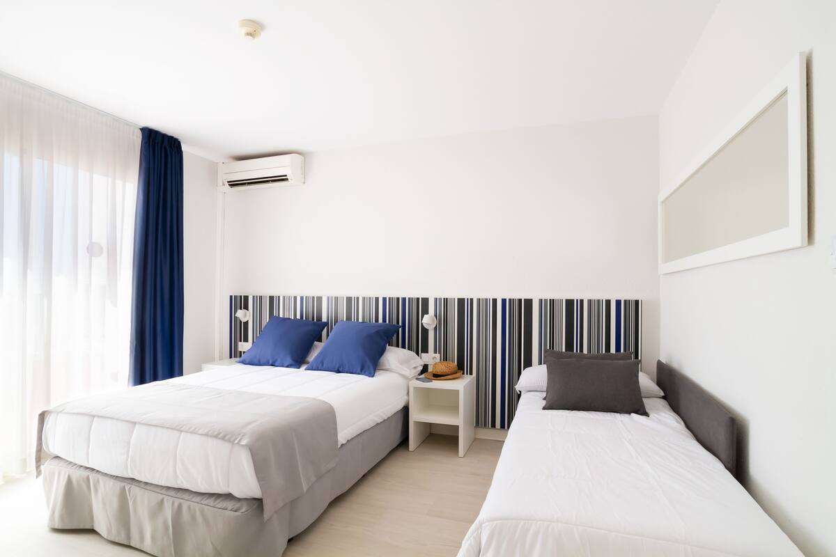 A hotel room with two beds, a striped headboard, a small bedside table with a lamp, and a large window with curtains.