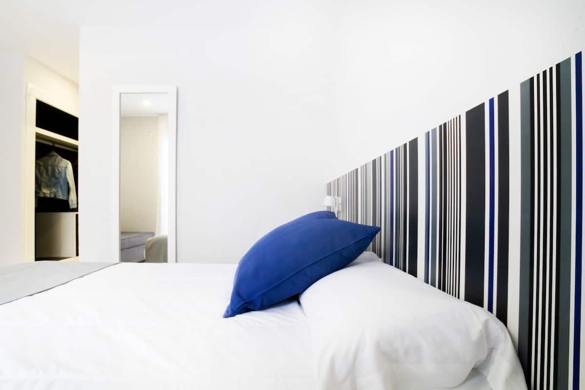 A modern hotel room with a bed, a blue pillow, a striped wall, and a closet with clothes.