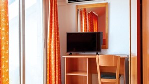 A room with a TV on a wooden stand, a chair, and a window with orange curtains. - Santa Susanna Resort Affiliated by FERGUS (Santa Susanna)
