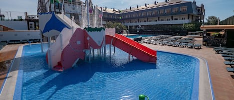 Una piscina con tobogán acuático y un tobogán rojo, rodeada de sillas de descanso y un edificio de hotel al fondo.