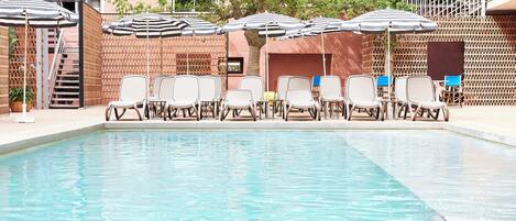 A pool area with lounge chairs and umbrellas, surrounded by a wooden fence and a building with a staircase.