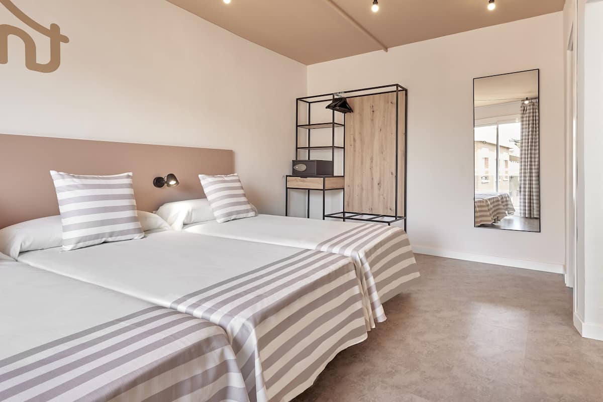 A modern hotel room with two beds, a wooden wardrobe, a mirror, and a view of the city.