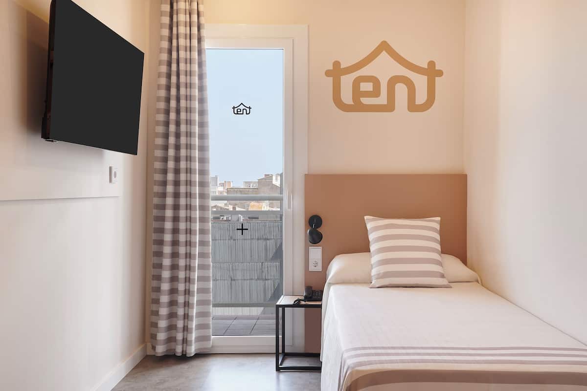 A hotel room with a bed, a TV mounted on the wall, a small table, and a view of the city through the window.
