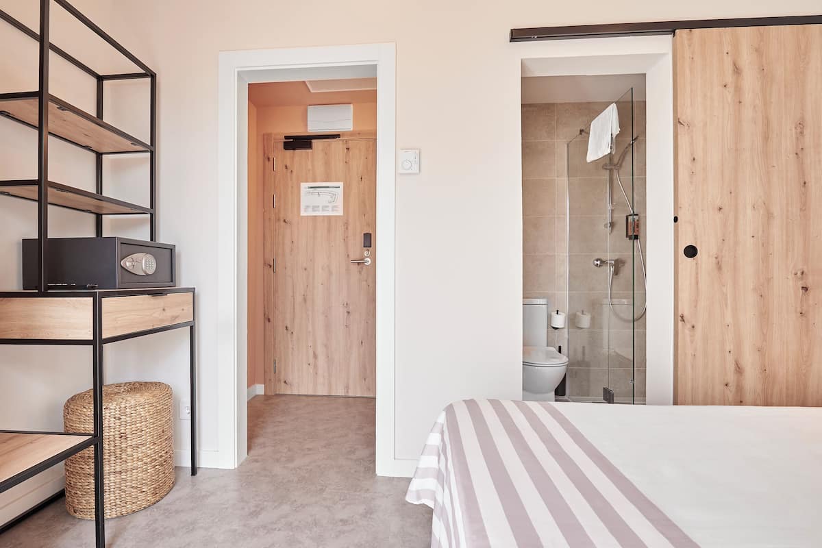 A bedroom with a bed, a bedside table, a radio, a wicker basket, and a wooden door leading to a bathroom with a shower.