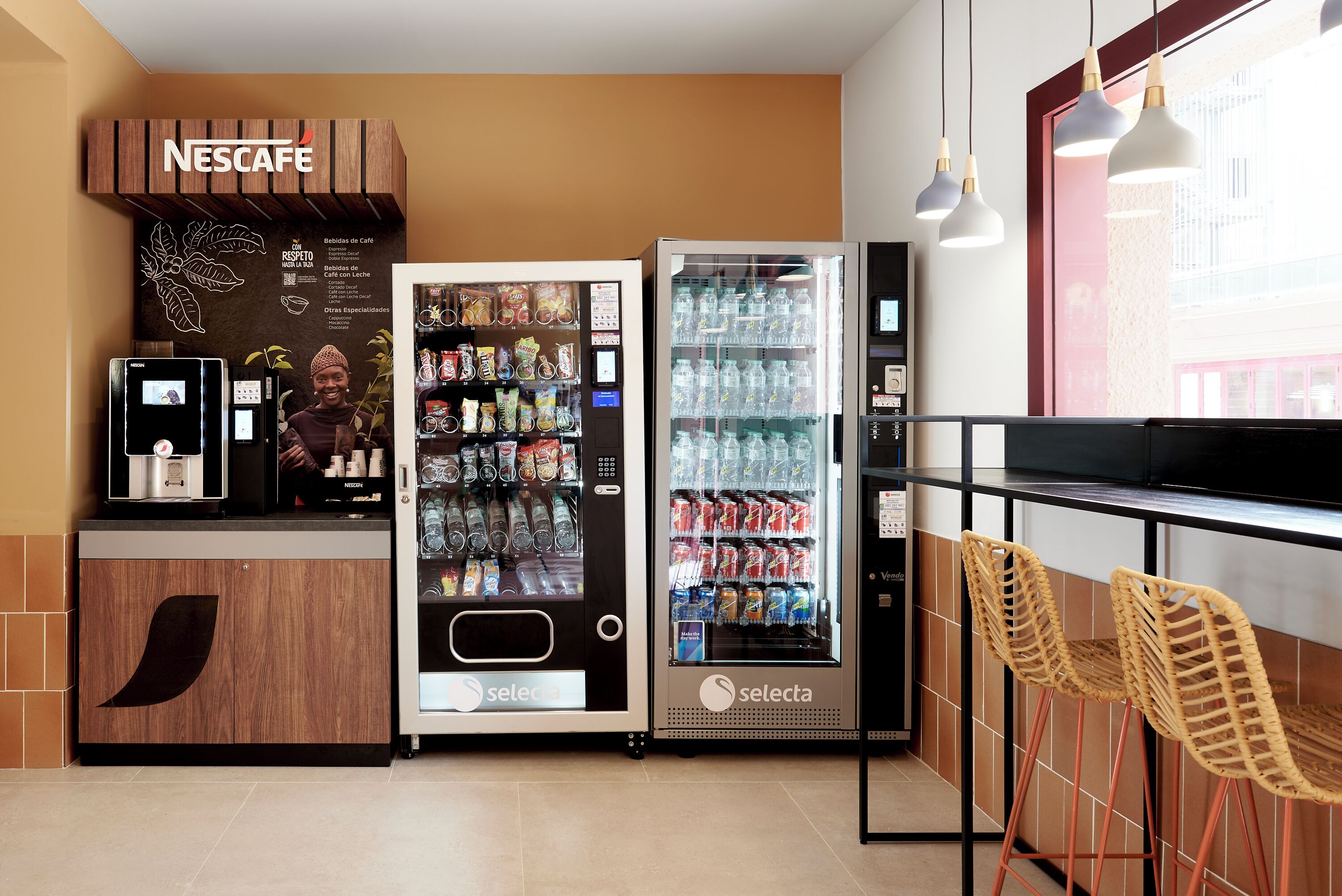 Ένας σταθμός καφέ Nescafe με αυτόματο πωλητή και ψυγείο, μια περιοχή με καθίσματα και καρέκλες από μπαμπού και ένα παράθυρο με θέα στα κτίρια.