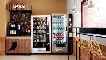 A Nescafe coffee station with a vending machine and a refrigerator, a seating area with wicker chairs, and a window with a view of buildings.