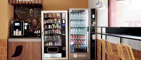 Una estación de café Nescafé con una máquina expendedora y un refrigerador, un área de descanso con sillas de mimbre y una ventana con vistas a edificios.