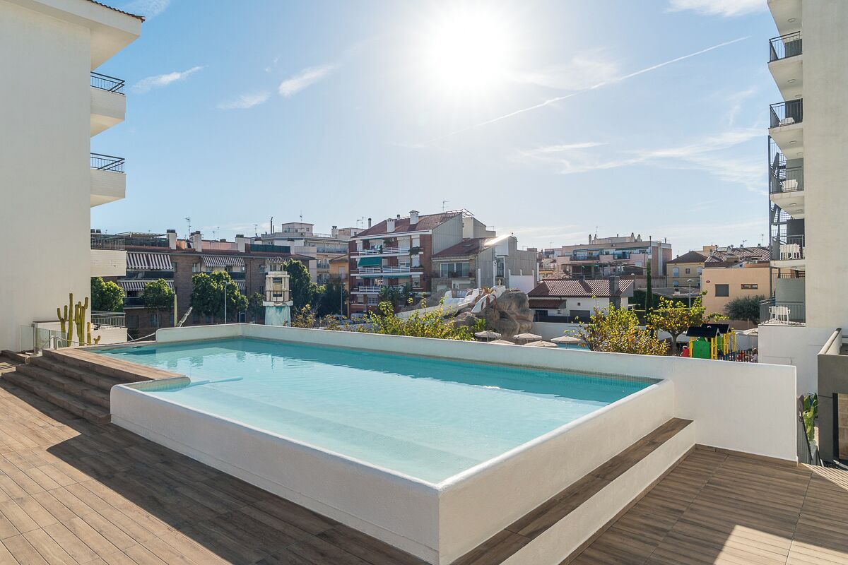 Une piscine rectangulaire avec une terrasse en bois, entourée de bâtiments et sous un ciel dégagé.
