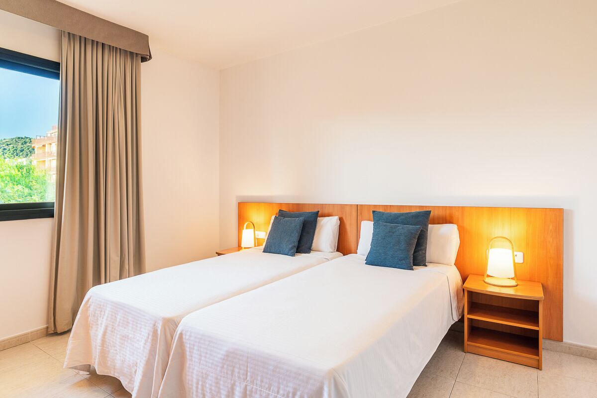 A hotel room with two beds, a nightstand with a lamp, and a window with curtains.