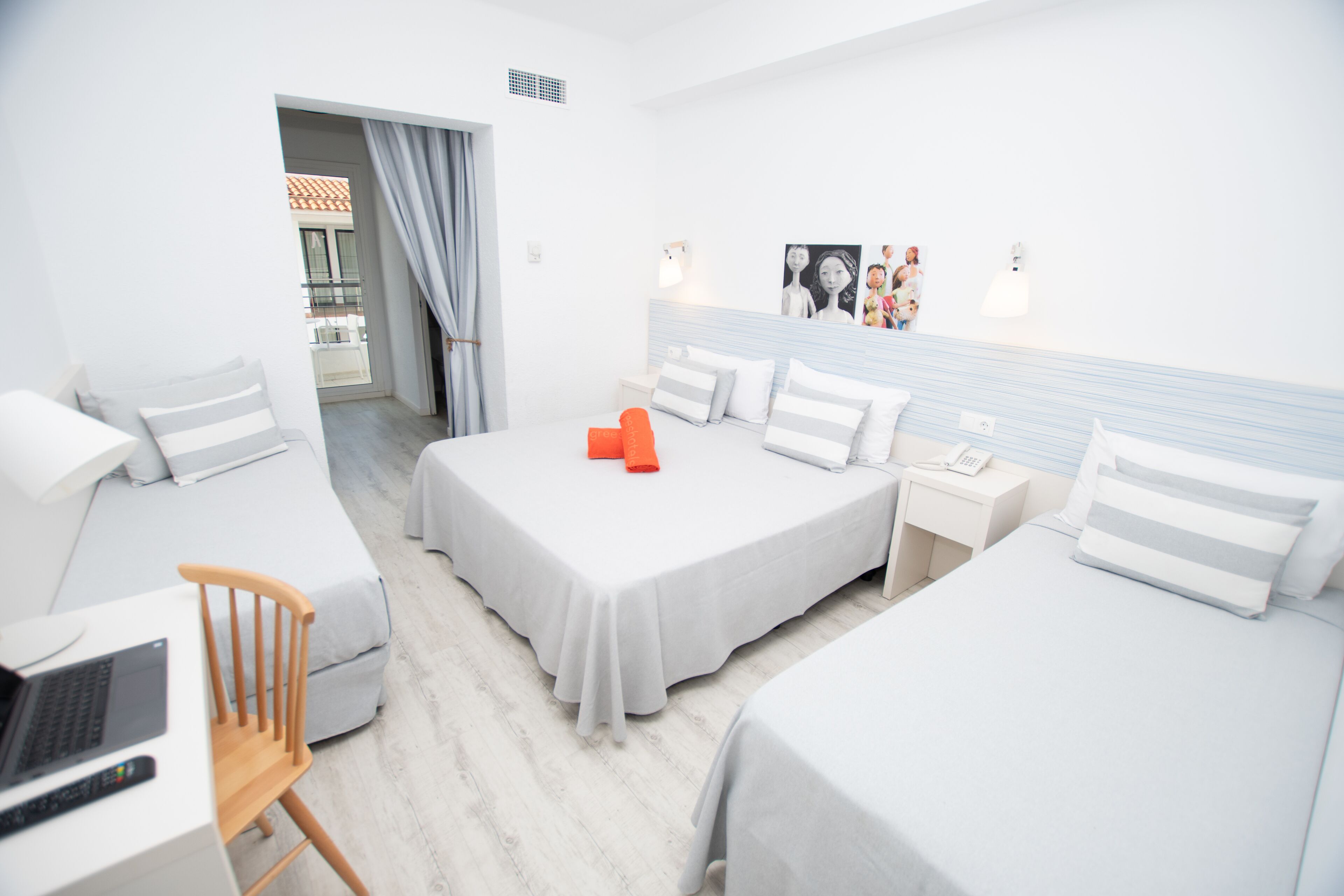 A hotel room with two beds, a desk, and a chair. There is a window with curtains, a bedside table with a lamp, and a wall with artwork.
