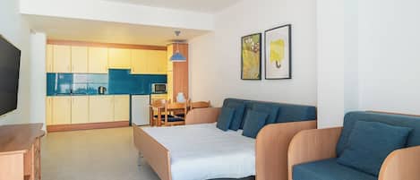 A modern hotel room with a bed, a sofa, a small table, a TV, and a kitchenette in the background.