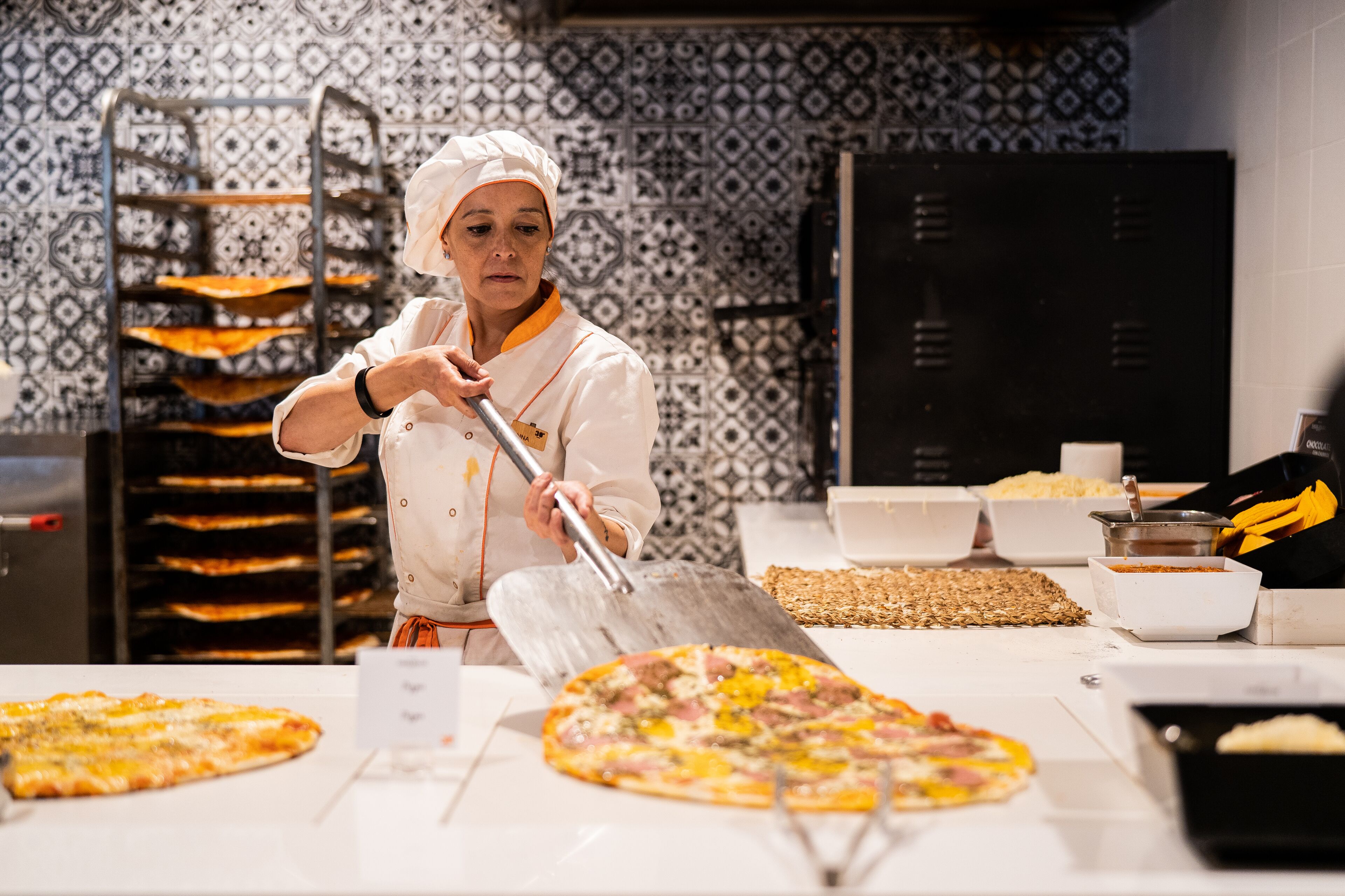 A chef preparing pizzas in a kitchen with patterned tiles and a rack of pizza pans.
