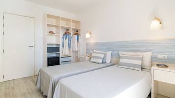 Habitación de hotel con dos camas, un armario con ropa colgada, una mesita de noche con una lámpara y un aplique de pared.