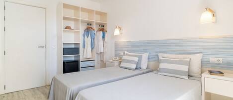 A hotel room with two beds, a wardrobe with hanging clothes, a bedside table with a lamp, and a wall-mounted light fixture.