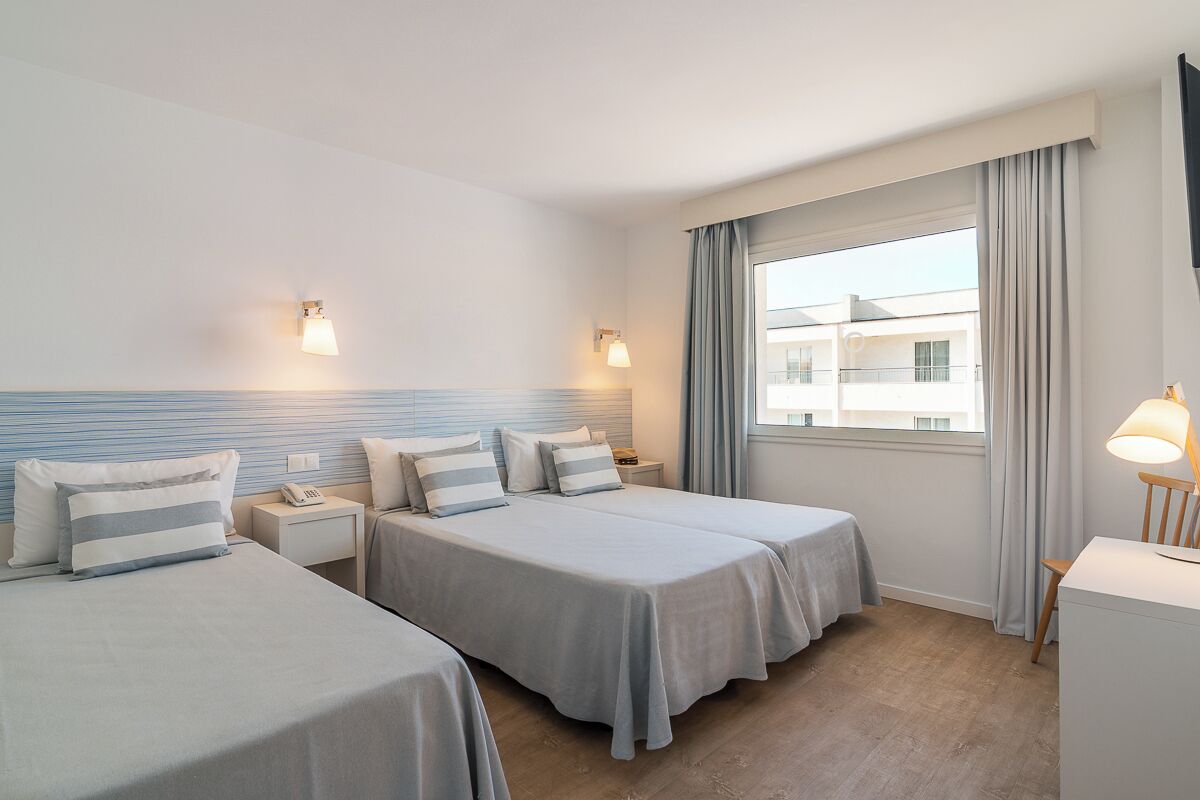 Habitación de hotel con dos camas, un escritorio, una silla, un ventanal con cortinas y dos lámparas fijadas a la pared.