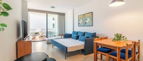 A modern, well-lit room with a bed, a sofa, a dining table with chairs, and a TV on a wooden cabinet.