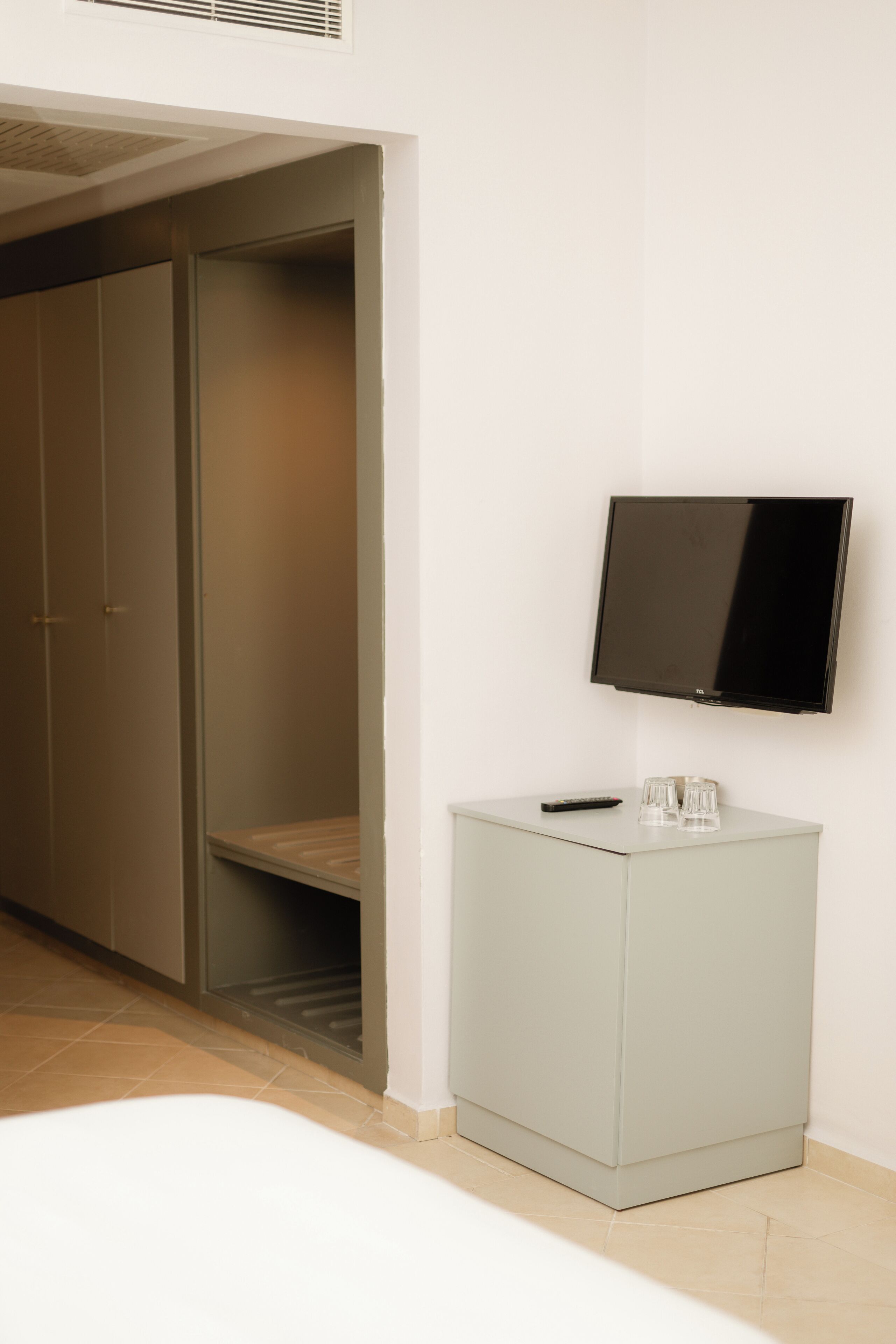 Una habitación con cama, televisor montado en la pared, armario y un mueble pequeño con vasos.