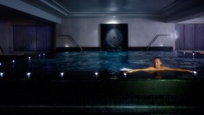 Una persona en una amplia piscina interior con un diseño moderno, que incluye una obra de arte geométrica en la pared y un techo minimalista.