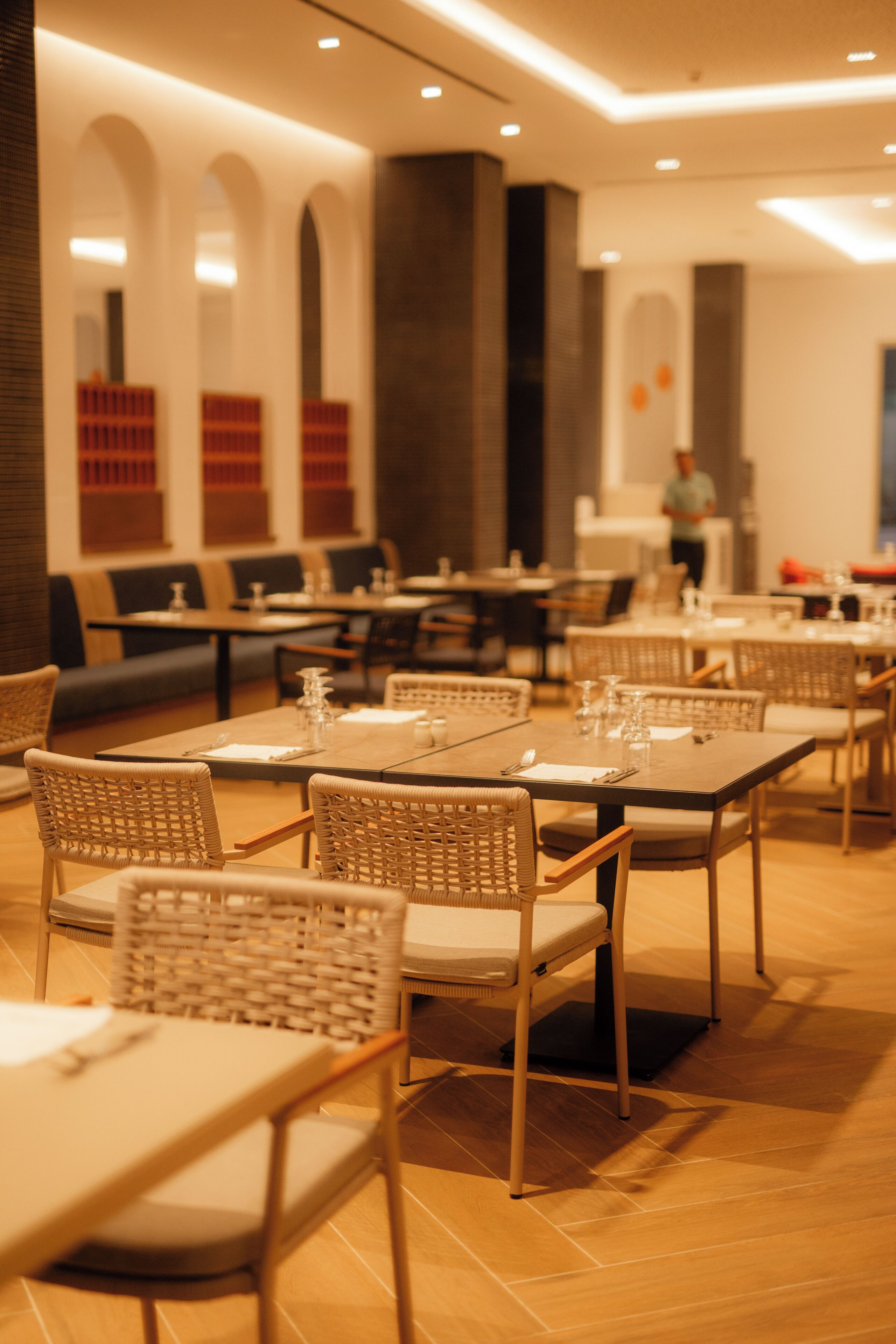 A restaurant with multiple tables and chairs, a bar area, and a person in the background.
