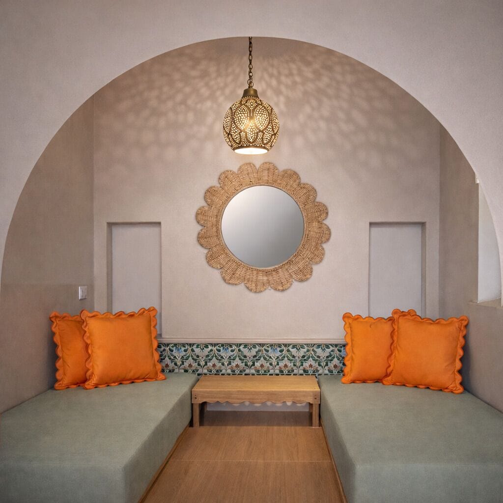 Una camera accogliente con una panca bassa in legno, due cuscini arancioni, uno specchio decorativo e un lampadario.