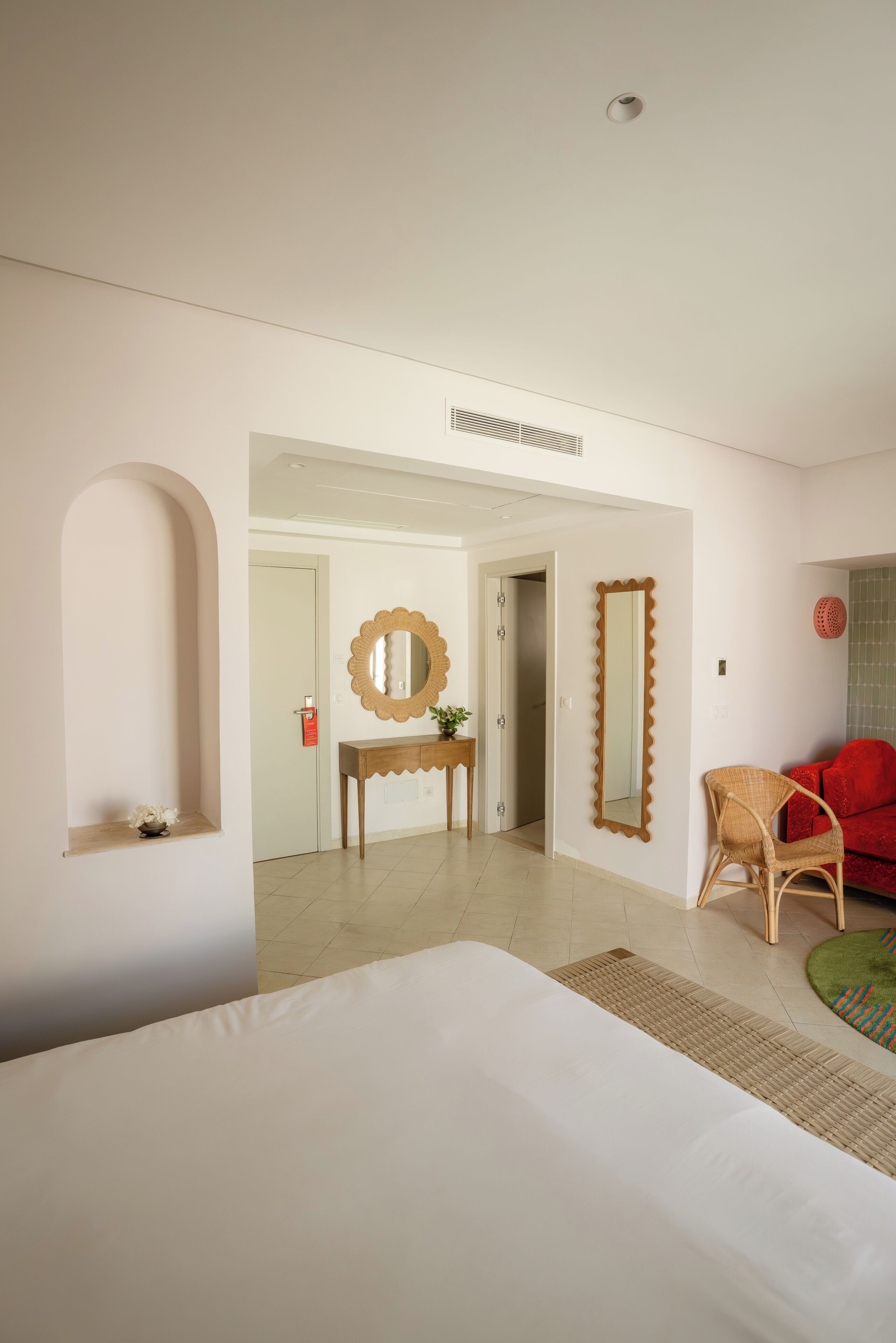 Une chambre d’hôtel avec un lit, un miroir, une chaise rouge, une console et une porte donnant sur une autre pièce.
