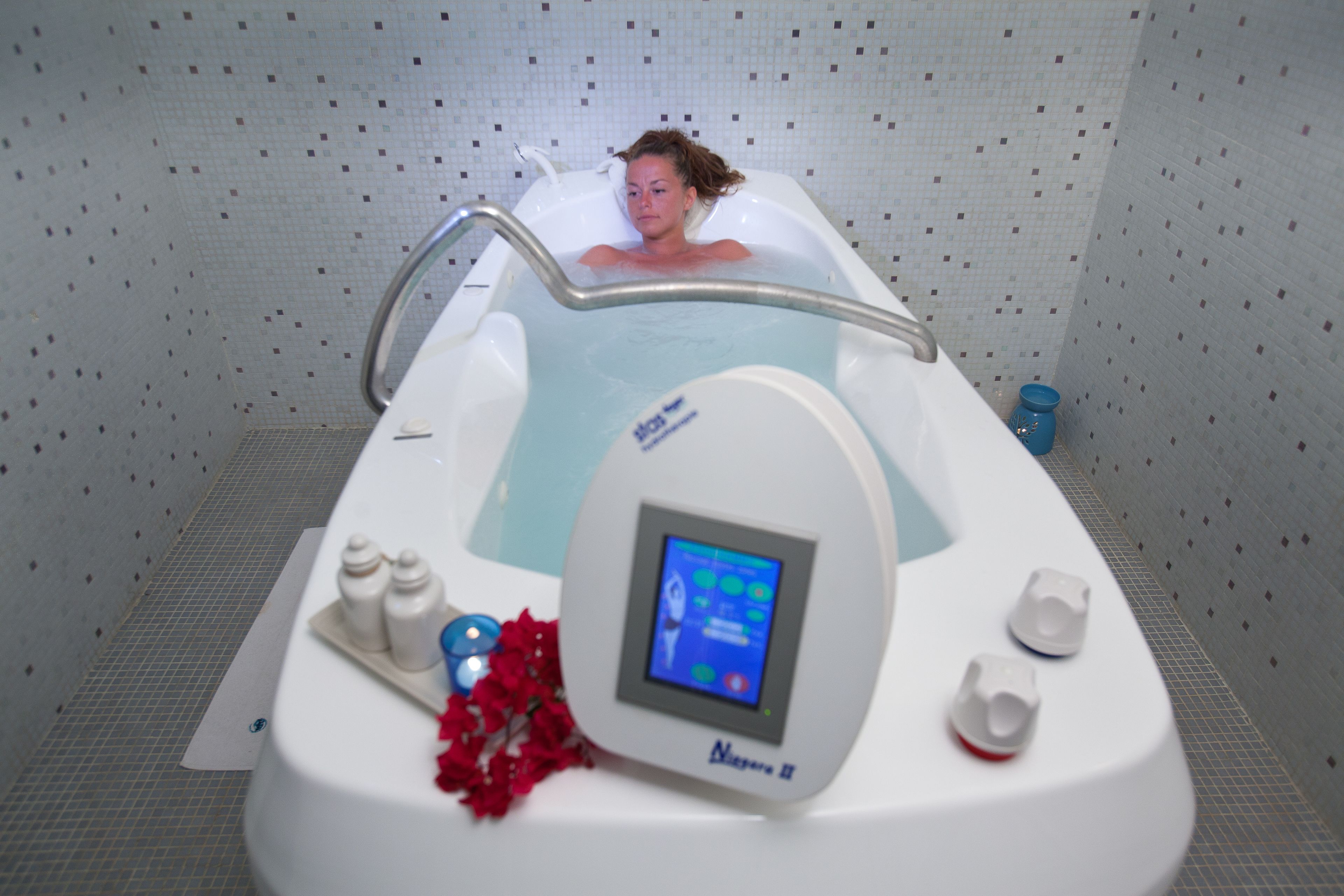 A spa bath with a control panel, a metal handrail, and a tiled wall.