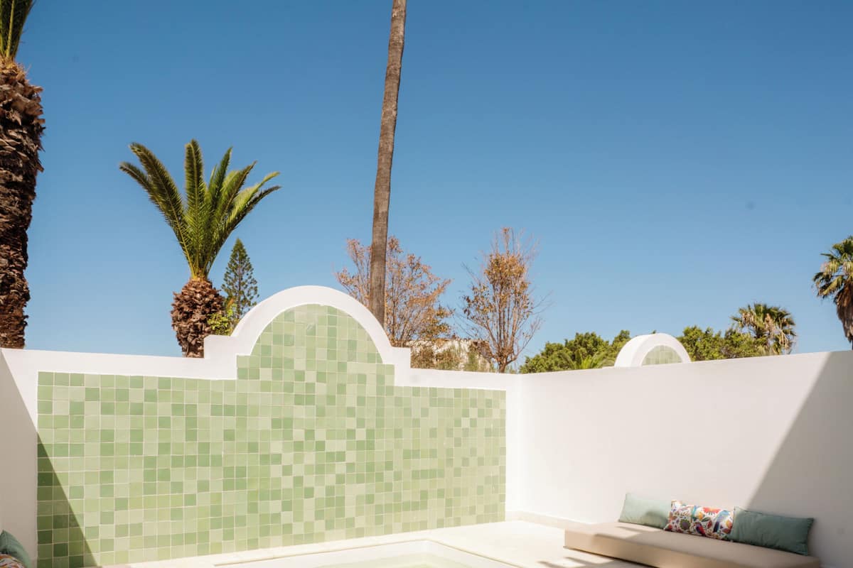 A pool area with a tiled wall, a seating area with cushions, and palm trees.