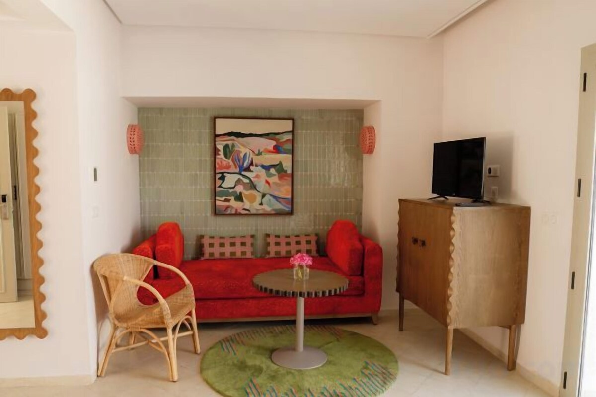 Una stanza con un divano rosso, un tavolino rotondo, una sedia, una televisione su un mobile e un quadro appeso al muro.