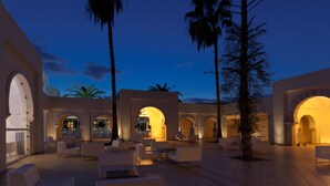 Un espacio al aire libre con mobiliario blanco, palmeras y un edificio con arcos.