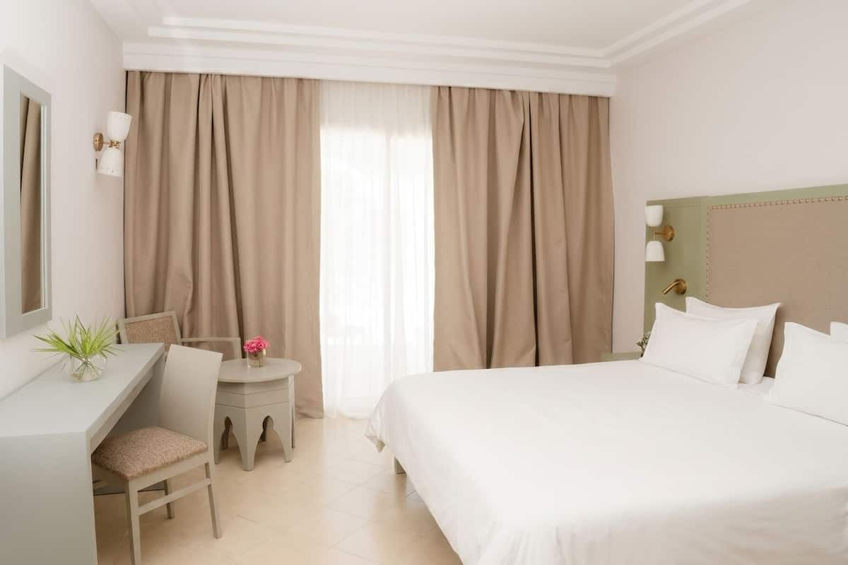 A hotel room with a bed, a desk, a chair, a small table with a plant, and a window with curtains.
