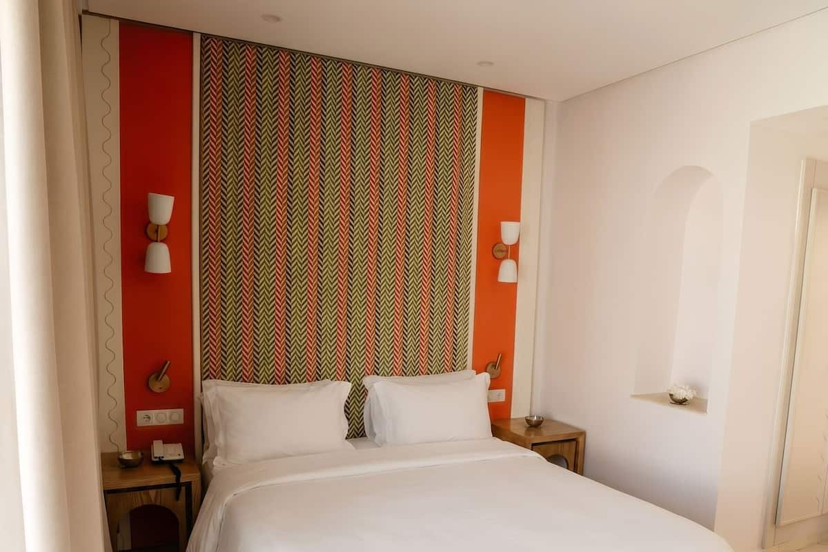 A hotel room with a bed, bedside tables, a bench, a patterned wall, and a door.
