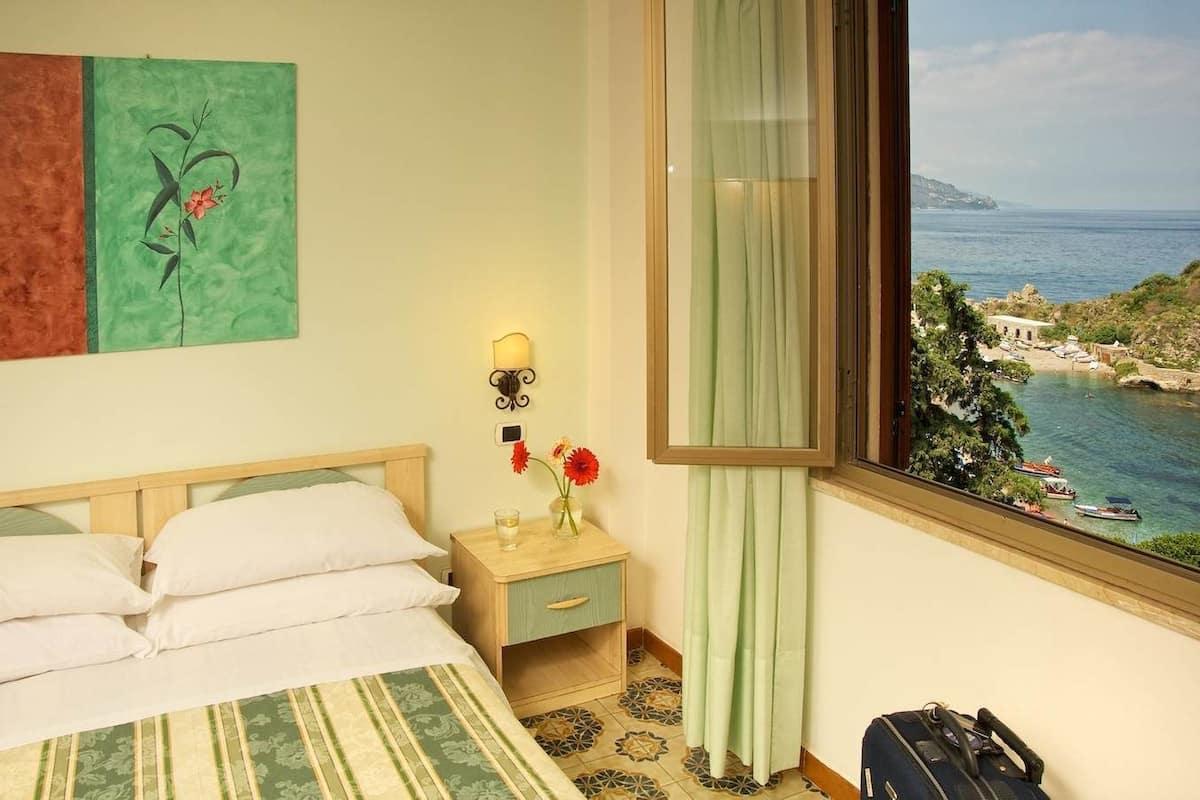 Une chambre d’hôtel avec un lit, une table de chevet, un vase de fleurs, une fenêtre donnant sur la côte et une valise posée sur le sol.