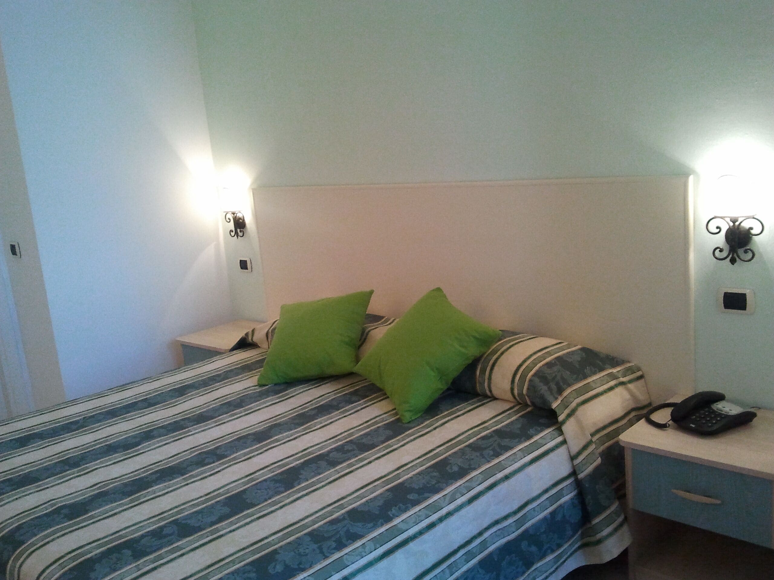Een bed met gestreept beddengoed en twee kussens, een nachtkastje met een telefoon, en wandlampen.